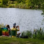 A serene scene of two individuals fishing by a lakeside, one seated on a colorful chair casting a line, while the other captures the tranquil moment with a camera, surrounded by lush greenery and calm waters.